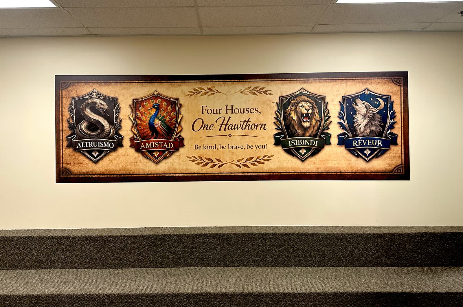 Four Houses, One Hawthorn identity mural installed above a stairwell at Hawthorn Academy. Four custom crests (Altruismo serpent, Amistad peacock, Isibindi lion, Reveuk wolf) with the tagline Be kind, be brave, be you.